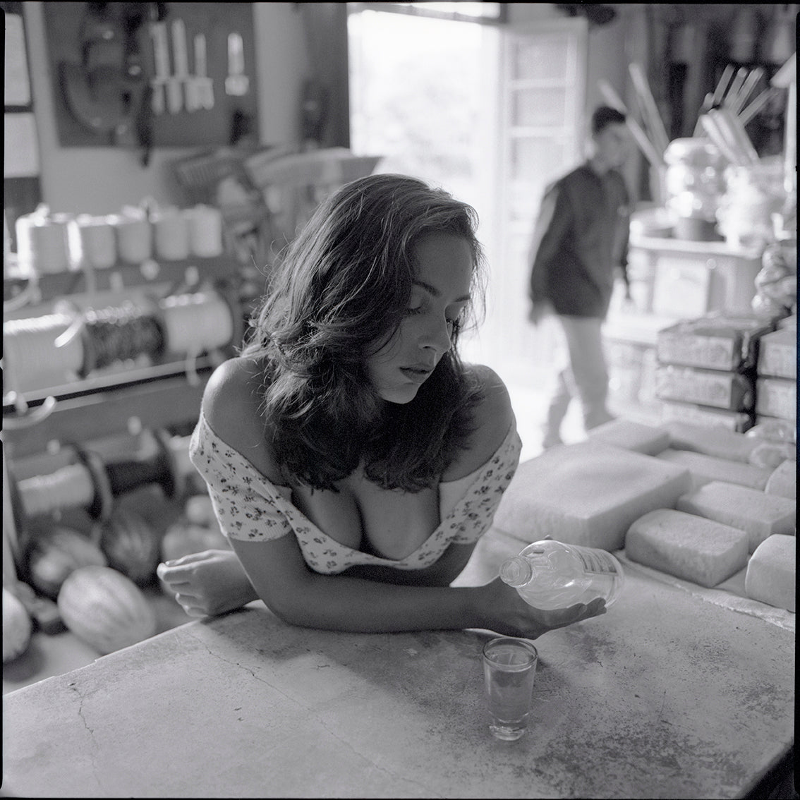 In the vintage counter of a rural general store, a young woman becomes part of the scene with quiet ease and natural grace. Around her, shelves hold the everyday life of the countryside — tools, bread, cheese, rope, soft drinks, and cachaça — while people come and go on the dusty streets outside. At times she drinks in silence, absorbed in her own thoughts; at others, she smiles softly, as if touched by a passing conversation, a memory, or the slow rhythm of the afternoon. Her presence does not seek attenti