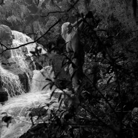 Analog black and white photograph, captured in 2004 and recently digitized. This original work reflects an intimate connection between the body and nature, where the female figure moves in the shadows as if searching for the light. The timeless texture of IlfordHP5 Plus film enhances the contrast, creating a mysterious atmosphere in which the body merges with the forest and a delicate waterfall in the background. Enigmatic, yet harmonious, the image evokes strength and serenity, balancing the human presence
