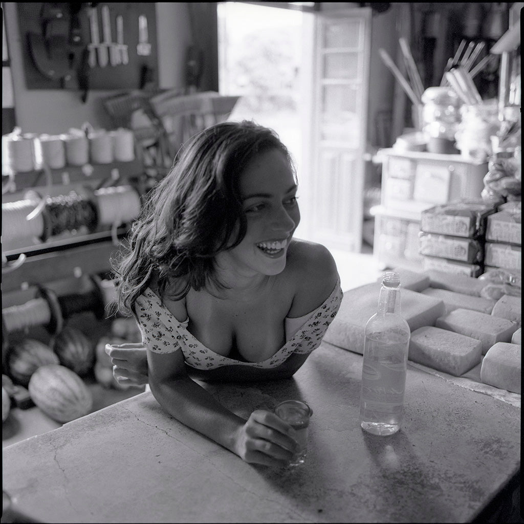 In the vintage counter of a rural general store, a young woman becomes part of the scene with quiet ease and natural grace. Around her, shelves hold the everyday life of the countryside — tools, bread, cheese, rope, soft drinks, and cachaça — while people come and go on the dusty streets outside. At times she drinks in silence, absorbed in her own thoughts; at others, she smiles softly, as if touched by a passing conversation, a memory, or the slow rhythm of the afternoon. Her presence does not seek attenti