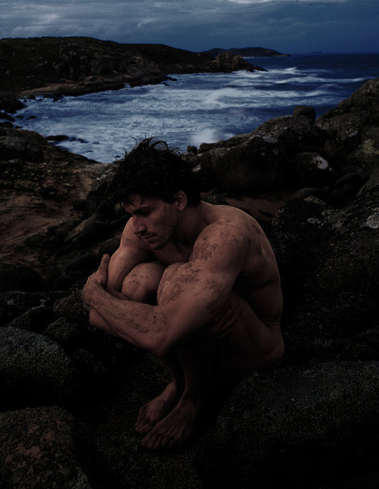 "Thinking Man," also part of the "Elemental" series, presents the same figure in the same rocky coastal landscape, now gathered into a more compact and introspective pose. With his hands and feet drawn close to the body as he rests upon the rocks, the man recalls the timeless resonance of Rodin’s The Thinker, yet here the gesture is displaced from monumentality into the raw openness of nature. The photograph transforms this familiar sculptural echo into a quieter meditation, in which the body appears both g